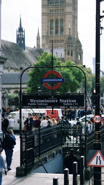 A busy street scene in Westminster with historic Gothic-style buildings, including the Palace of Westminster with its prominent clock tower. In the foreground, a sidewalk and roadway are visible with
